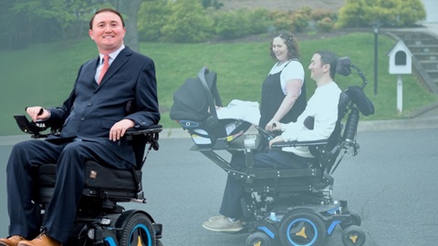From Joshs Perspective: A man in a suit uses a power wheelchair and smiles at the camera. In the background, he is with a woman and a baby in a car seat, all smiling as he moves his wheelchair along a sunny street.