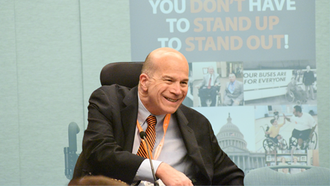 A man in a suit and striped tie, seated in a wheelchair, smiles during an event. Behind him is a display with photos and the message: YOU DONT HAVE TO STAND UP TO STAND OUT!—capturing Kents Perspective.