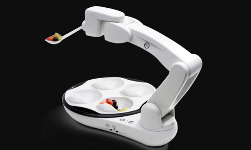 A robotic feeding arm holds a spoon with food above a segmented plate containing small portions of various foods, against a black background. This innovative device showcases specialty products designed for mealtime assistance.