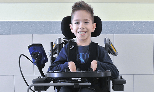 A young pediatric boy with short brown hair smiles while sitting in a power wheelchair, using alternative access controls. A tablet is mounted to the side of his chair against a light-colored tiled wall.
