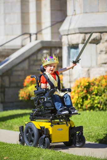 A young child wearing a crown and costume sits in a yellow complex power wheelchair, holding a toy sword aloft with excitement outside near a stone building and bright flowers.