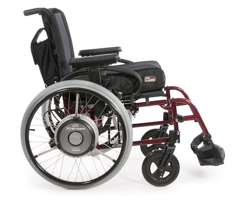 A side view of a manual wheelchair with large rear wheels, small front caster wheels, padded seat, armrests, footrests, and a maroon frame, featuring a Power Assist add-on motor attachment on the rear wheel.