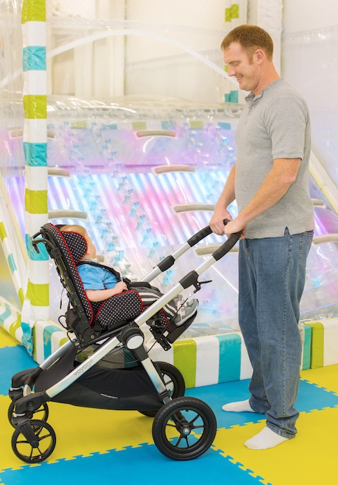 A man in a gray shirt and blue jeans stands on a colorful foam mat, smiling at a baby seated in a stroller. In the background, there is a brightly lit, pastel-colored play structure.