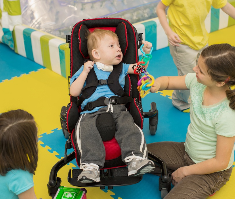 A young child in a supportive wheelchair is playing with colorful toys held by another child, while two more children sit nearby on a vibrant foam mat. They are all interacting in a playful, inclusive environment.