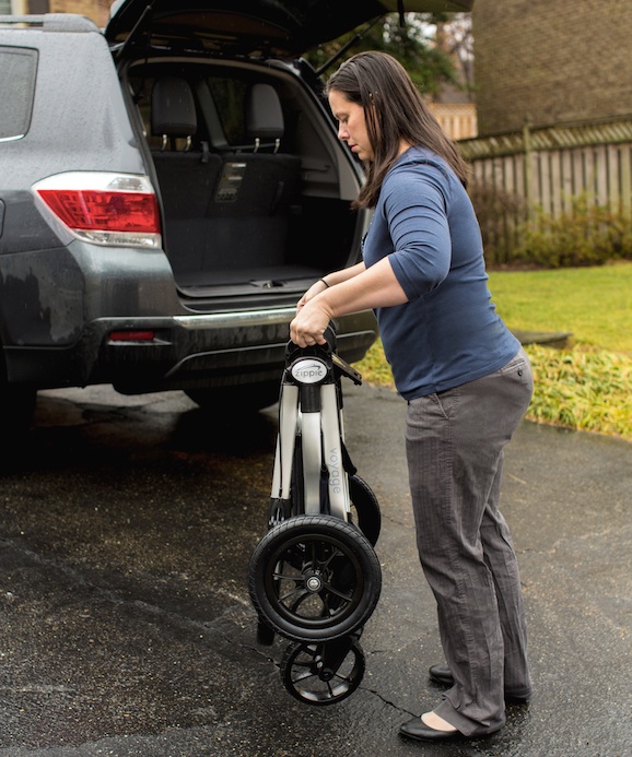 A woman stands on a driveway, folding a stroller near the open trunk of an SUV, ready to load it into the vehicle. In the background, there’s a residential yard with trees and a fence.