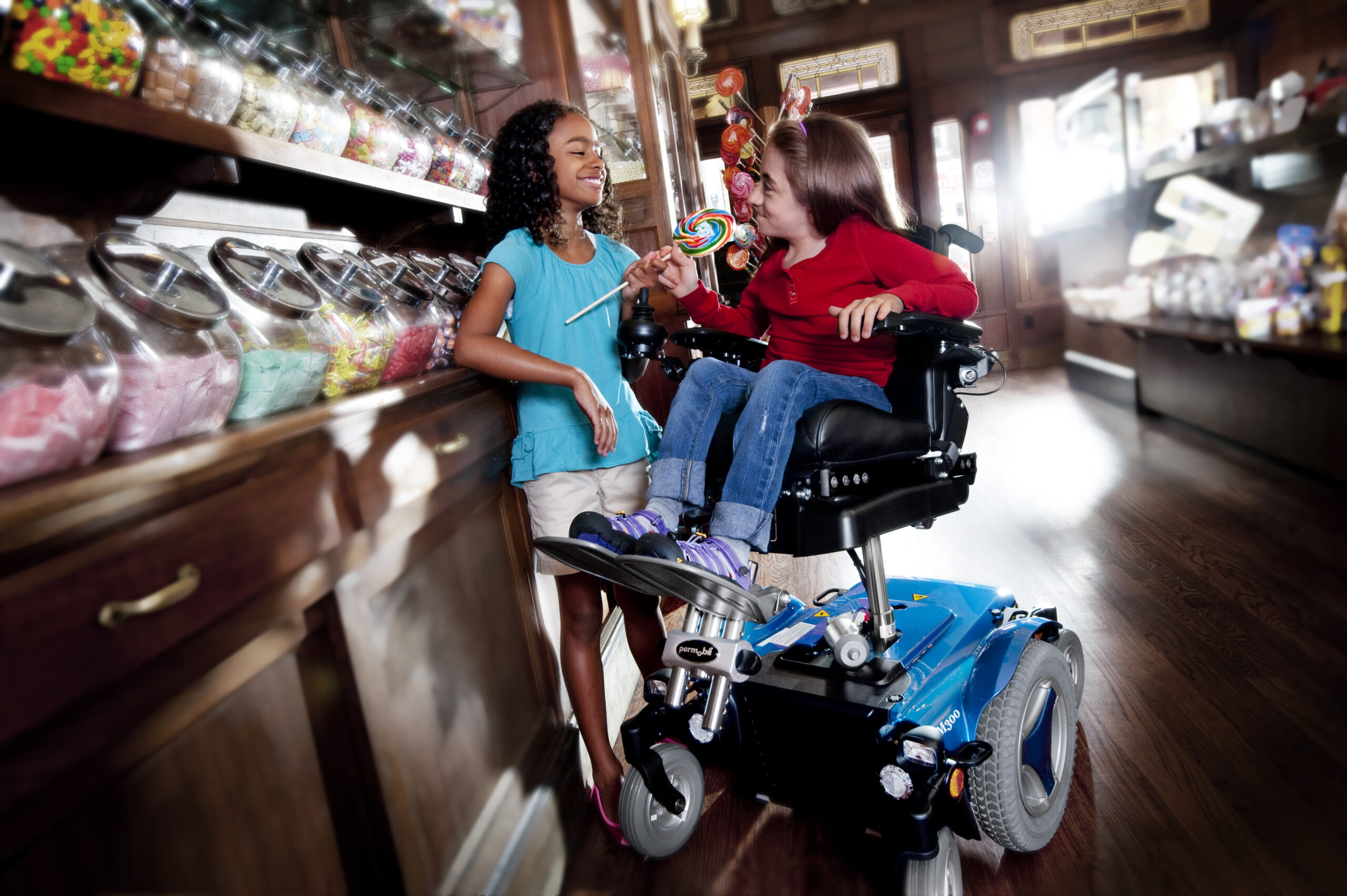 Two girls in a candy store smile at each other. One stands holding a lollipop while the other, seated in a Complex Power Tilt Wheelchair, holds a colorful lollipop. Glass jars filled with sweets line the wooden shelves beside them.