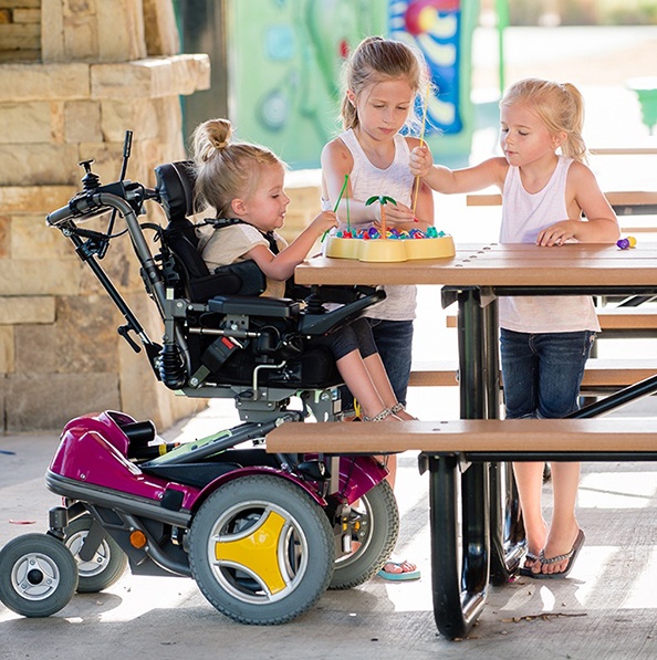 Three young girls, one using a complex power wheelchair, play together at a picnic table with colorful toys. They are outdoors, smiling and engaged in the activity.