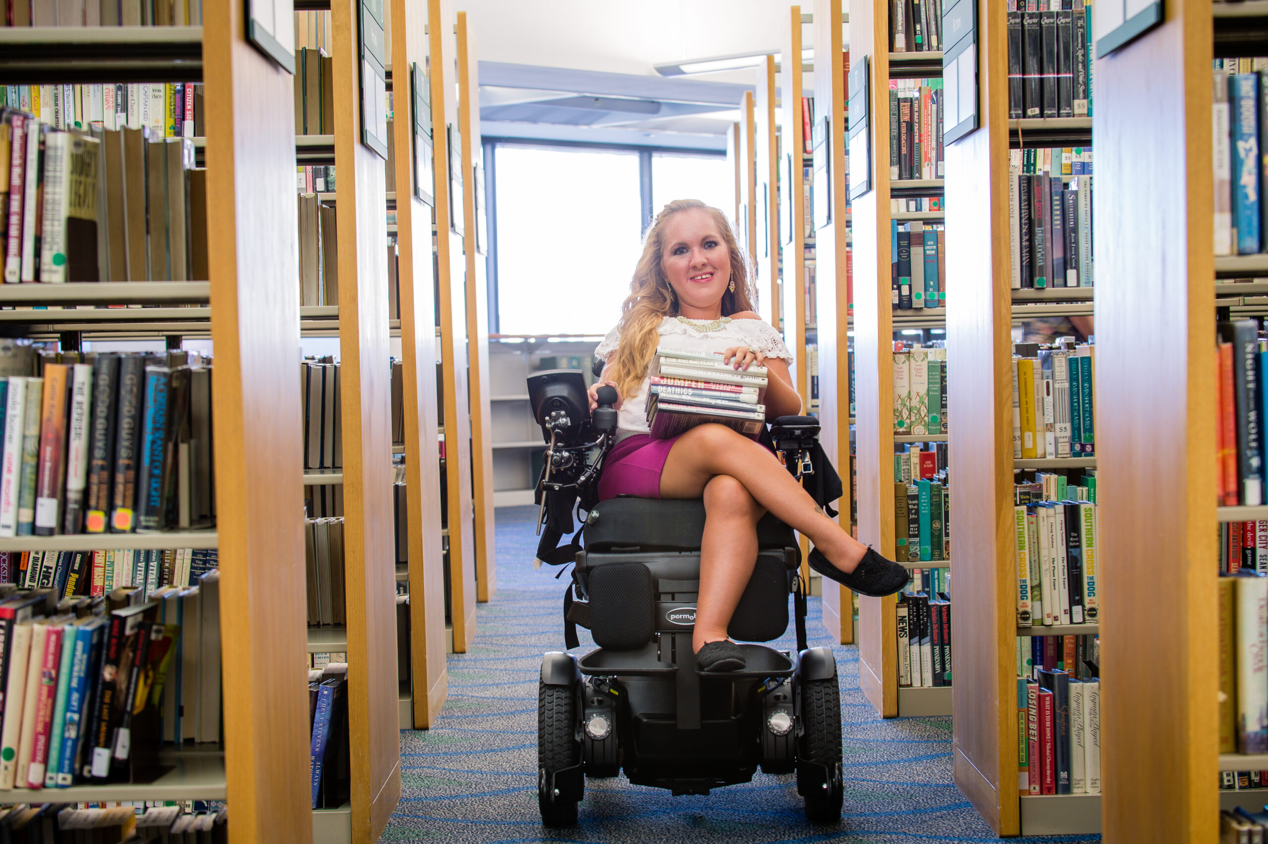A woman in a complex power wheelchair holds a stack of books and smiles while sitting between bookshelves in a brightly lit library.