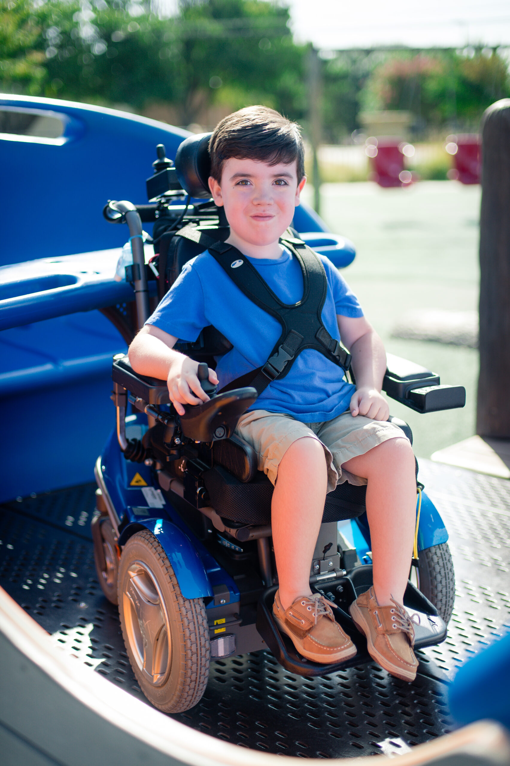A young boy with short brown hair, wearing a blue shirt and khaki shorts, sits in a pediatric power wheelchair at an outdoor playground, smiling at the camera.