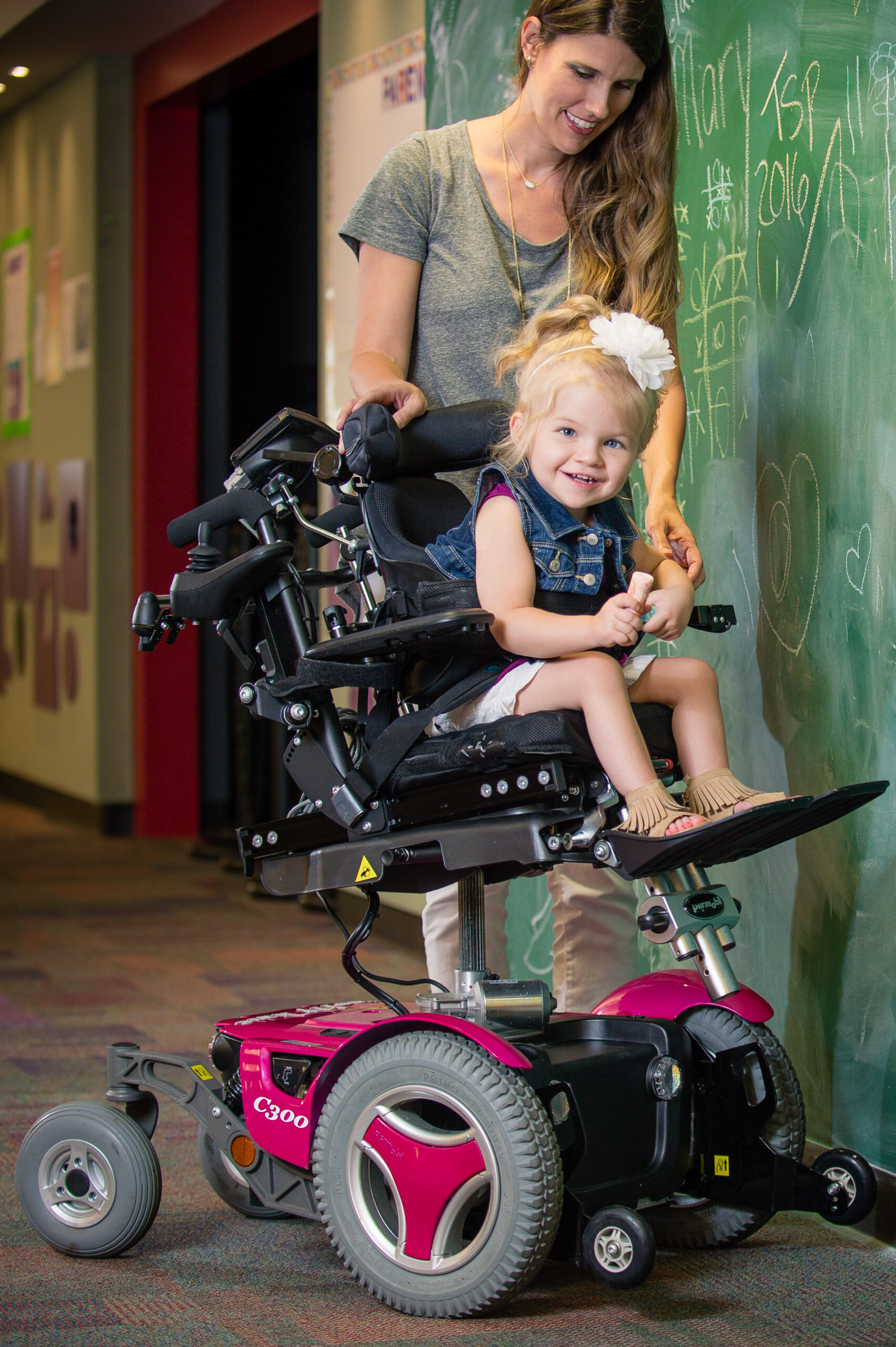 A smiling young girl with a white bow in her hair sits in a pink power wheelchair with Power Seat Elevators, while a woman stands behind her. Both look happy in a colorful indoor setting with a chalkboard filled with drawings.
