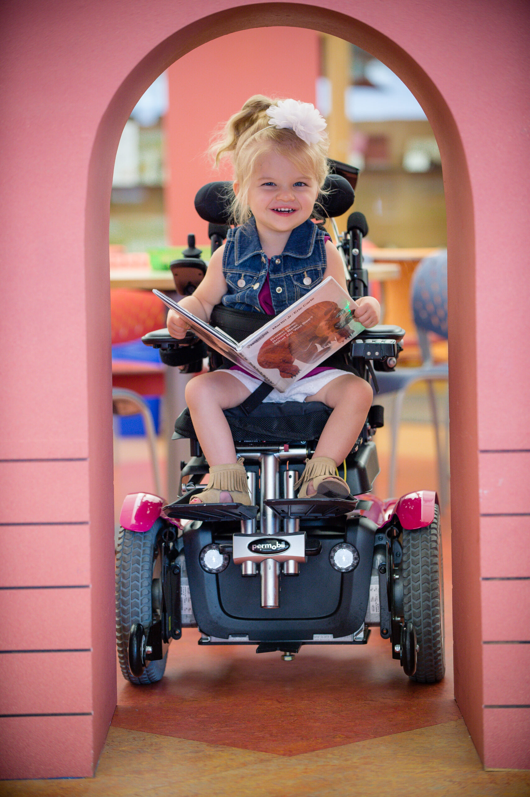 A young girl in a denim dress and white hair bow smiles as she holds an open book under a pink archway, seated in a pediatric wheelchair. The setting appears to be a library or classroom.