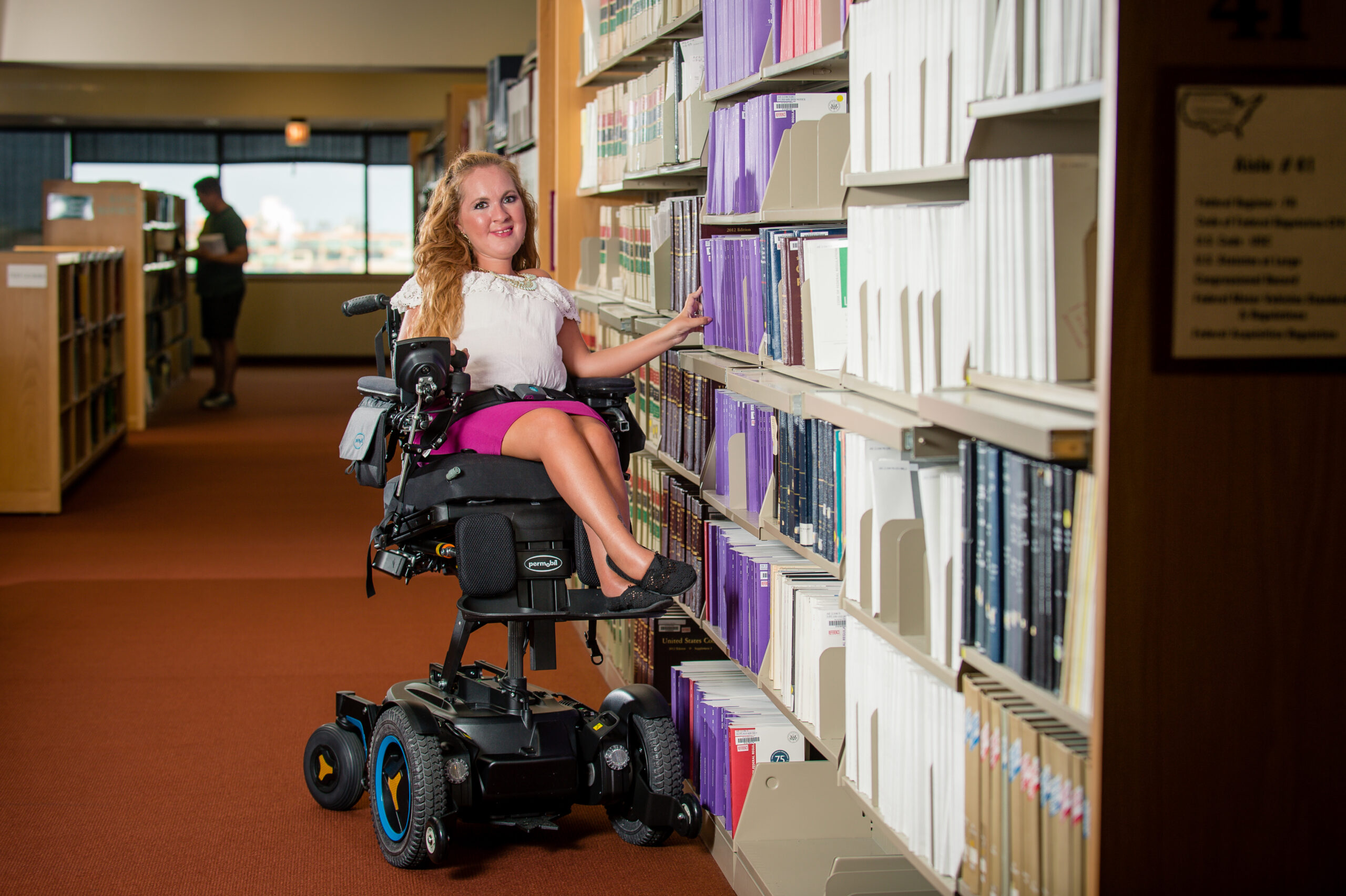 A woman in a motorized wheelchair with Power Seat Elevators smiles while reaching for a book on a library shelf, surrounded by neatly organized books and warm lighting.