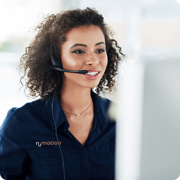 A woman with curly hair wearing a headset and a dark blue Numotion shirt is smiling while looking at a computer screen, ready to assist customers with their order timeline.