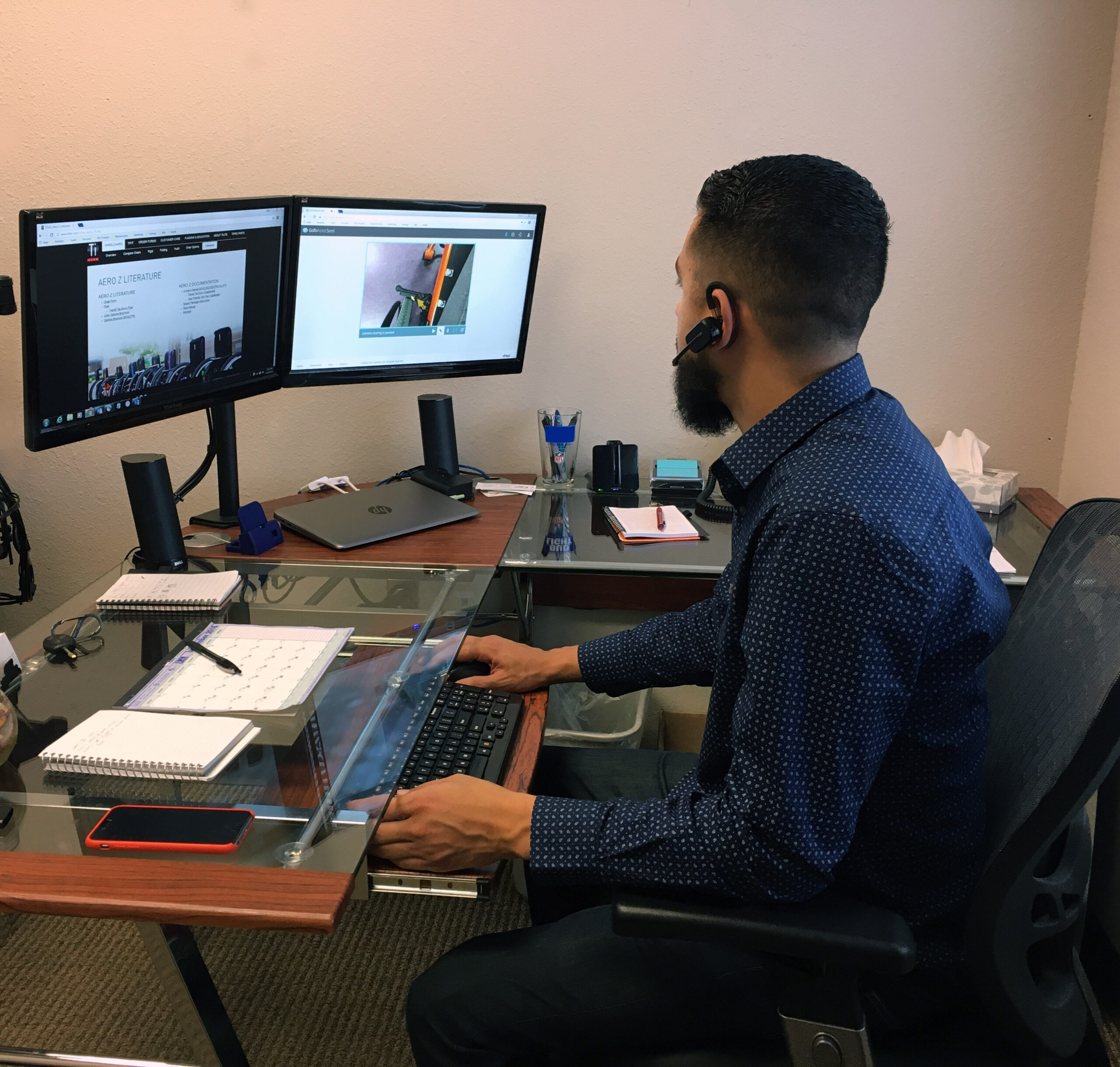 A man with a beard, wearing a blue shirt and headset, sits at a glass desk working on a computer with two monitors, providing remote service. Office supplies, notebooks, and a smartphone are on the desk.