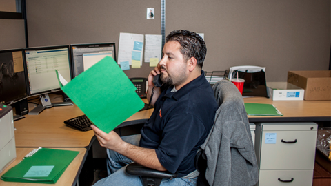 A man sits at a desk in an office, holding a green folder and talking on the phone about Support. Computer monitors, paperwork, and office supplies are visible around him.