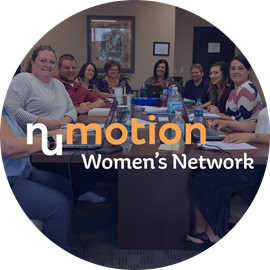 A group of women and men sit around a conference table with laptops and papers. Text overlay reads Numotion Womens Network, an Employee Resource Group. They are smiling in a professional office setting.