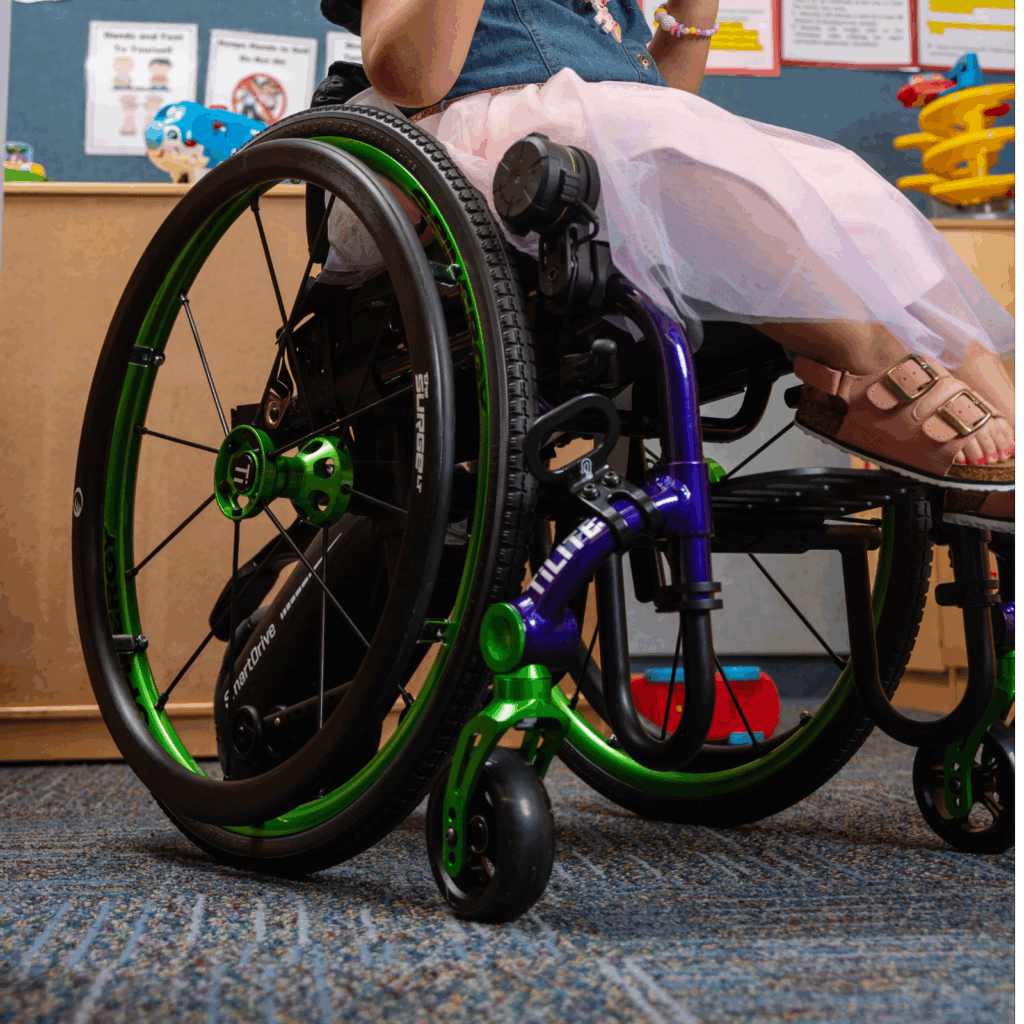 A young child wearing a pink dress and sandals sits in a green and black pediatric wheelchair with add-on power assist, on a carpeted classroom floor, colorful toys and classroom materials visible in the background.