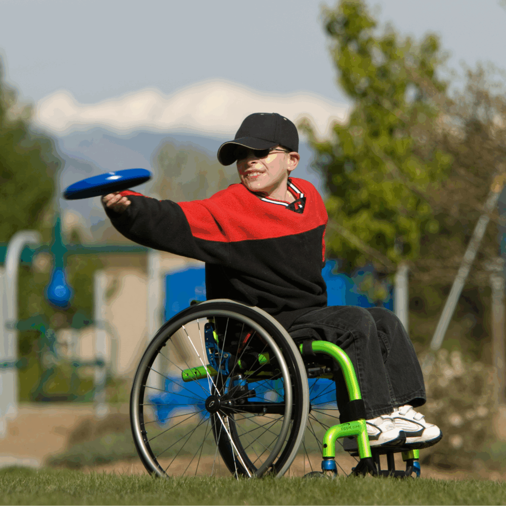 A boy in an ultra-light wheelchair smiles while playing with a blue frisbee outdoors on a grassy field, with playground equipment and snow-capped mountains visible in the background.
