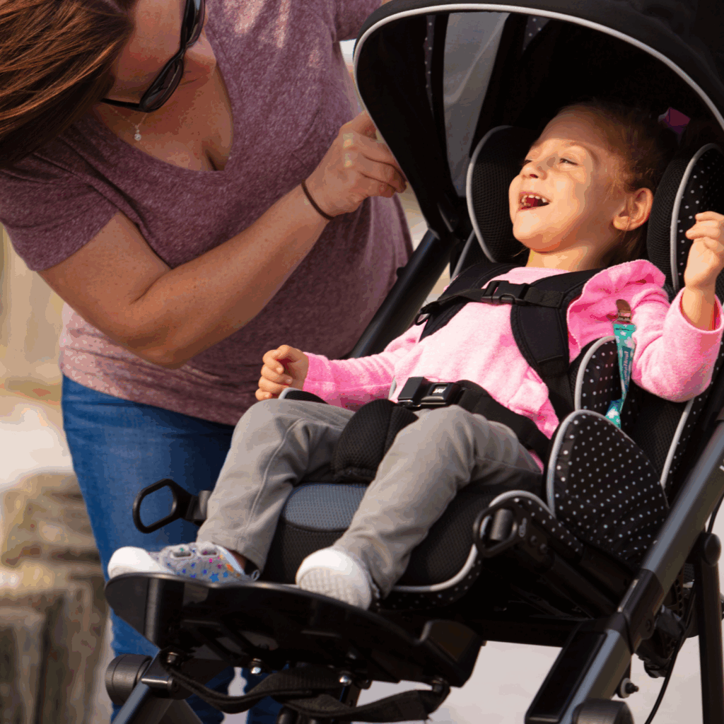A woman in sunglasses adjusts the harness of a smiling young girl in a wheelchair stroller outdoors. The girl, wearing a pink sweater and gray pants, appears happy and comfortable in her adaptive stroller.