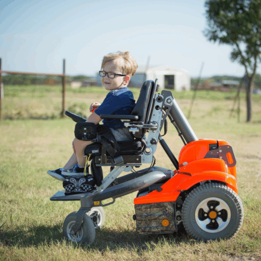 A young boy with glasses smiles while sitting in a pediatric power wheelchair on the grass, with a barn and trees in the background on a sunny day.