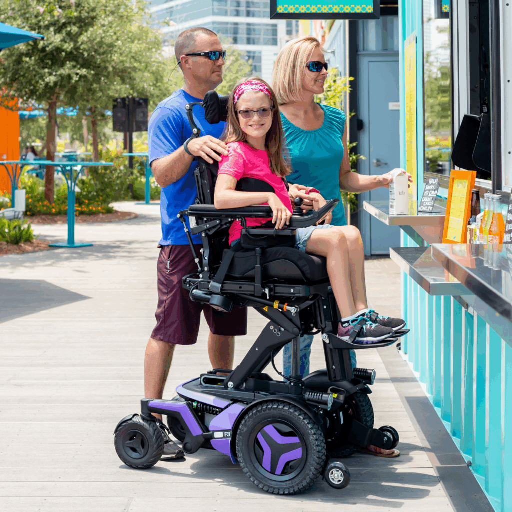 A young girl using a motorized wheelchair with Power Seat Elevators and purple accents sits at an outdoor counter with two adults, all smiling in sunglasses. The bright, sunny scene features lush greenery in the background.