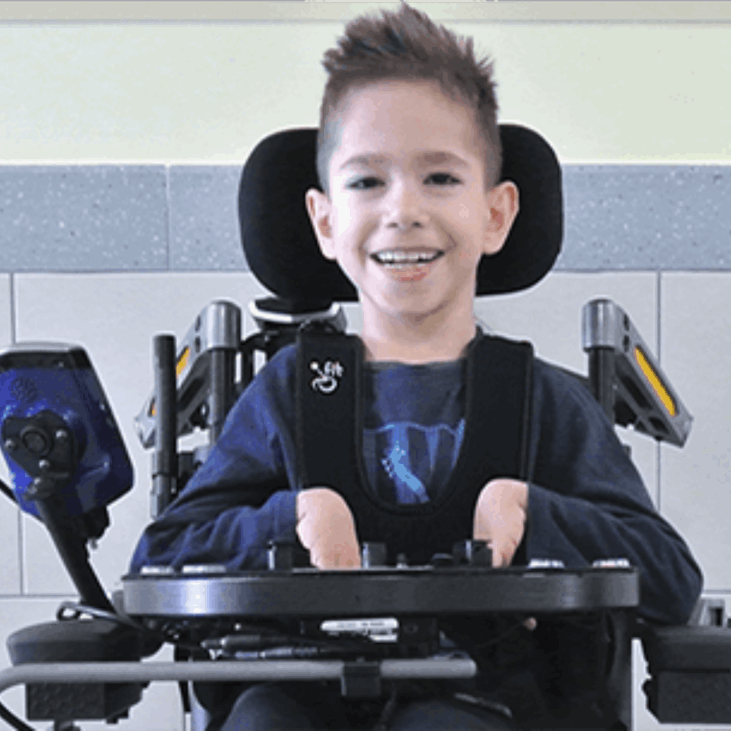 A young pediatric boy with short brown hair sits in a powered wheelchair, smiling at the camera. He wears a dark shirt and uses adaptive equipment with alternative access controls, including a tablet mounted to the side.