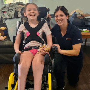 A smiling young girl using a pediatric mobility device holds a toy cheetah, while an adult woman in a navy shirt kneels beside her, also smiling. They are indoors, with toys and furniture in the background.