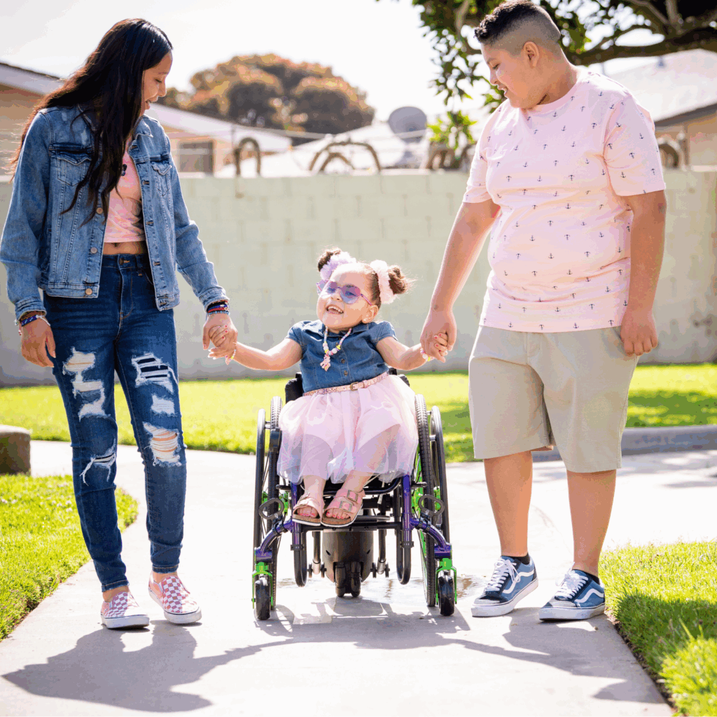 A young girl using an ultra-light wheelchair smiles while holding hands with a woman and a boy as they walk outdoors on a sunny day. All three appear happy and casually dressed.
