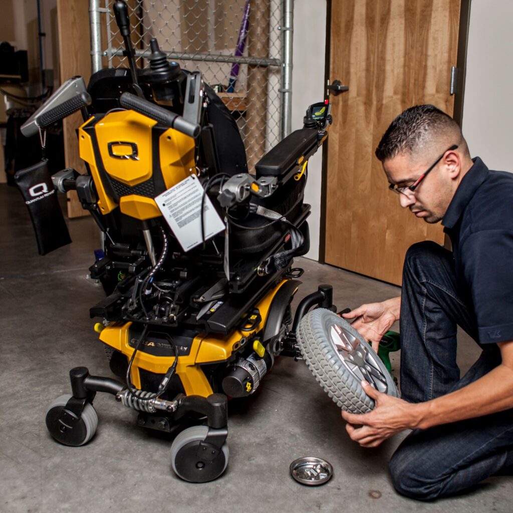 A man kneels beside a yellow and black power wheelchair in an indoor workshop, performing a repair or inspecting one of its large rear wheels as part of in-shop service.