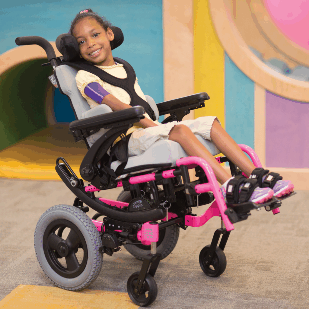 A young girl smiles while sitting in a pink and black pediatric tilt wheelchair indoors, wearing a yellow shirt and khaki shorts. The colorful background adds to the cheerful atmosphere.