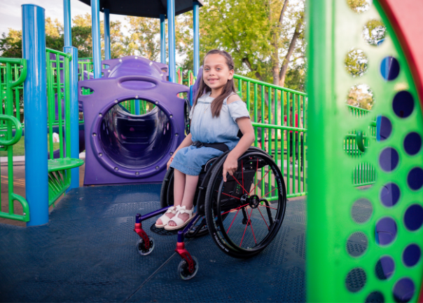 A young girl using pediatric mobility solutions smiles while sitting on an accessible playground surrounded by vibrant green and purple play structures. Trees and sunshine are visible in the background.