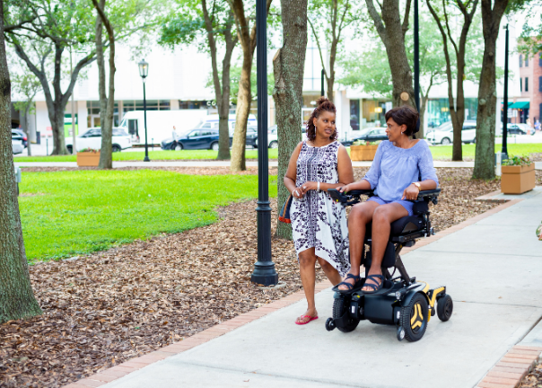 Two women enjoy a conversation outdoors; one is walking while the other uses a motorized wheelchair on a park-like sidewalk, offering a glimpse of what to expect in accessible outdoor settings with trees and grass.