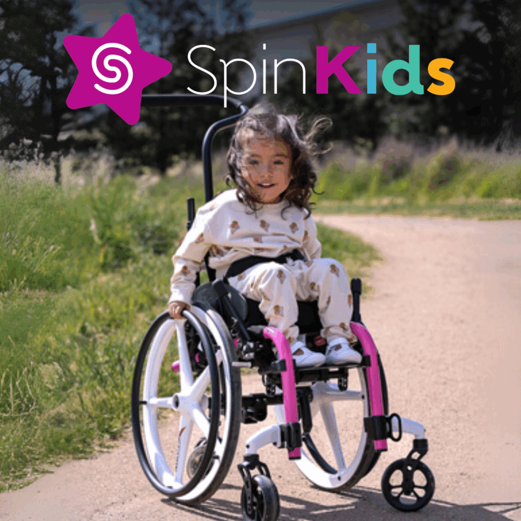 A young child in a pink and white pediatric mobility device smiles outdoors on a dirt path, with grass and trees in the background. The SpinKids logo with a purple star appears in the top left corner.