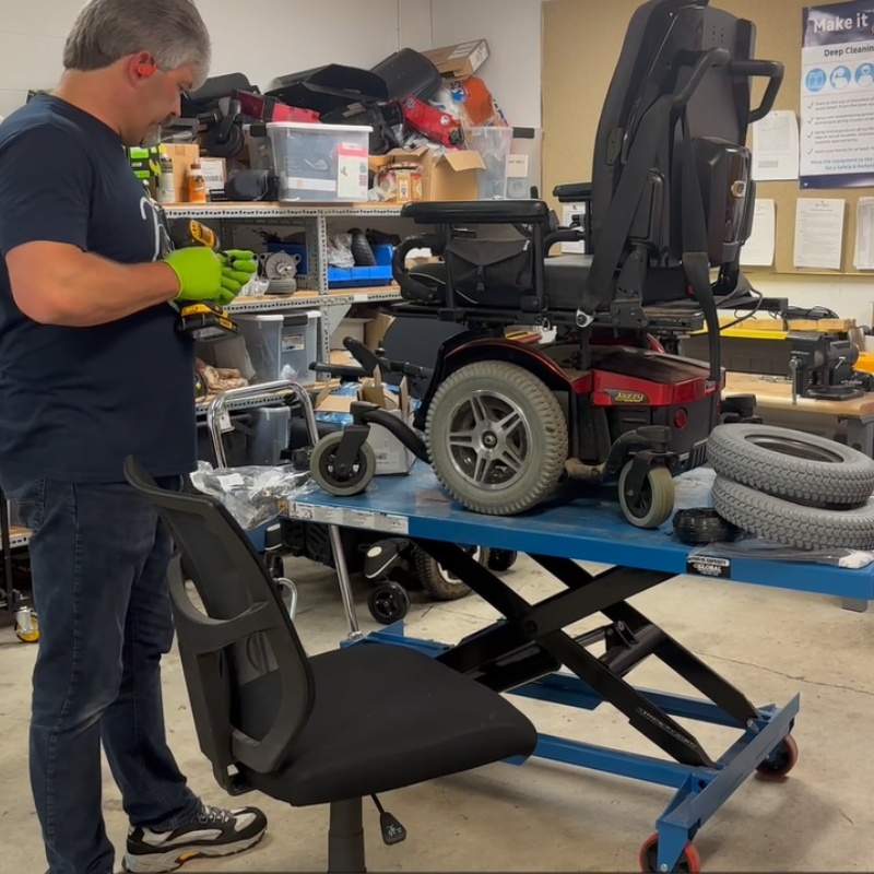 A man wearing gloves uses tools to repair a motorized wheelchair on a blue lift table in a workshop filled with equipment and supplies. An office chair sits nearby, highlighting the in-shop service environment.