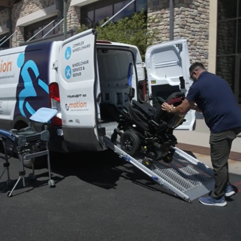 A man loads a power wheelchair into a white van labeled for Repair and In-Shop Service. Equipment is set up nearby, and the outdoor scene is next to a building with stone walls.