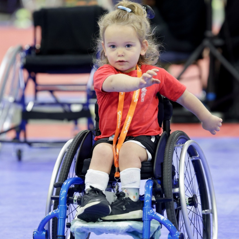 A young child with light brown hair in a ponytail, wearing a red shirt and white socks, sits in a blue wheelchair on an indoor court, reaching forward with one arm extended while playing.
