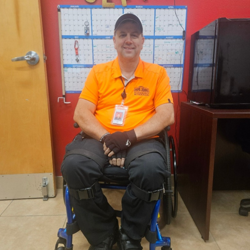 A man in an orange polo shirt and black pants sits in a wheelchair, smiling at the camera. He wears a cap, black gloves, and an ID badge, seated in an office with a calendar on the red wall behind him.