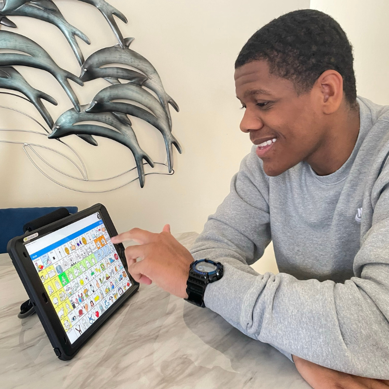 A young man sits at a table, smiling and using an assistive communication tablet with colorful picture symbols. Metal dolphin sculptures are mounted on the wall behind him.