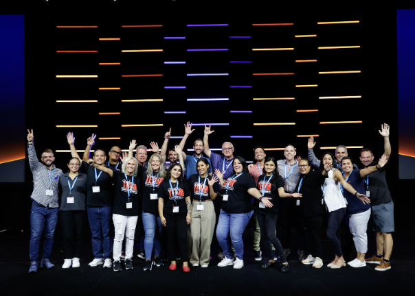 A group of people wearing conference badges pose and smile energetically on stage, with some raising their arms in excitement—giving you a glimpse of what to expect at this lively event. The background features a dark wall with colorful horizontal light strips.