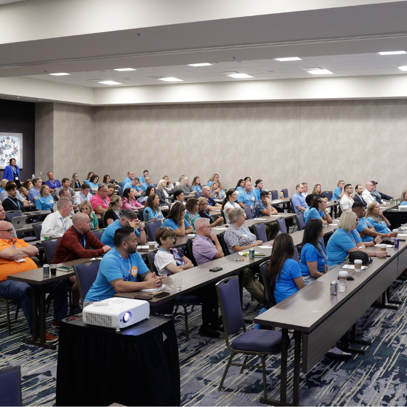 A large group of medical professionals sit attentively in rows of tables in a conference room, many wearing blue shirts, facing forward toward an unseen speaker or presentation. A projector sits at the front of the room.