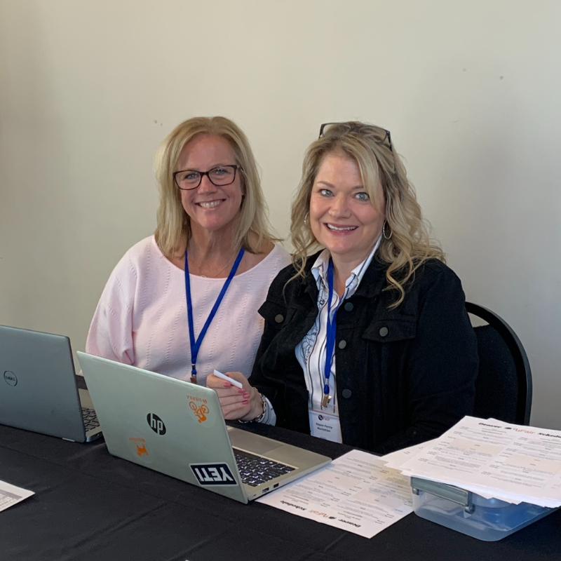 Two women with blonde hair, wearing glasses and blue lanyards, sit at a table with laptops and paperwork, smiling at the camera in a well-lit indoor setting, appearing to be medical professionals.
