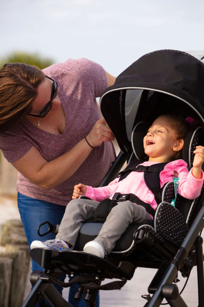 A woman smiles and leans toward a laughing young girl seated in a stroller. The girl, wearing a pink sweater and gray pants, enjoys the moment as the woman adjusts the stroller’s canopy outdoors.