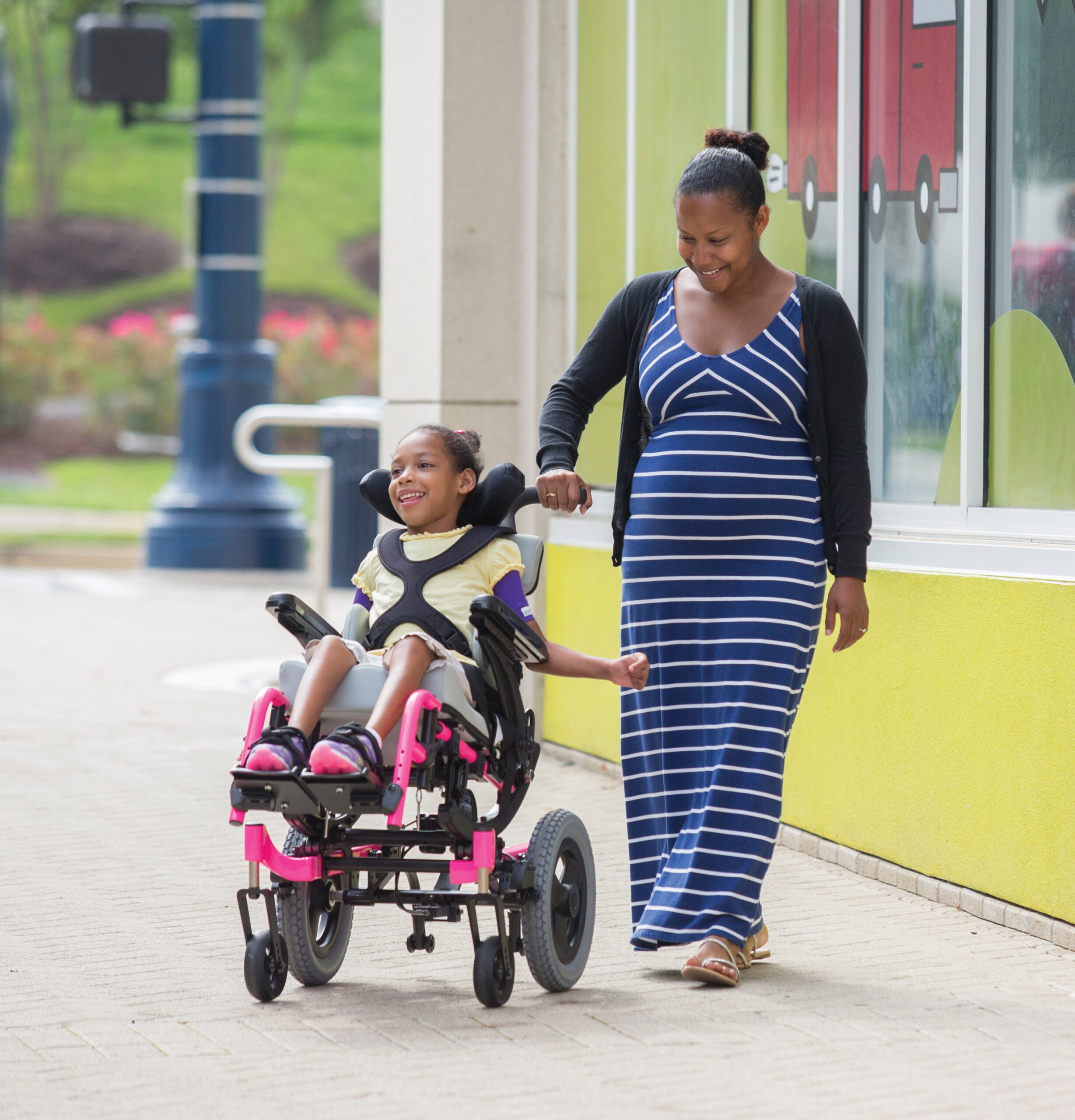 A smiling girl in a pink pediatric wheelchair moves along a sidewalk next to a woman in a blue and white striped dress, who is holding her hand. They appear happy, passing by a bright building with colorful windows.