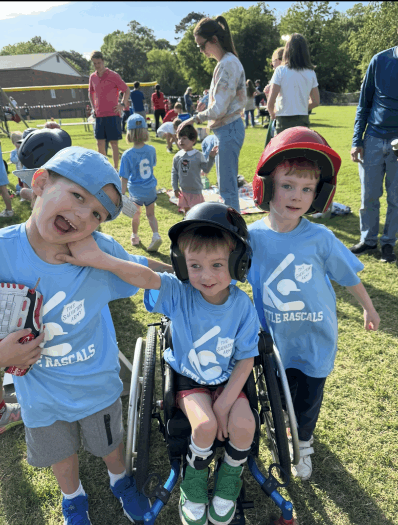 Three young children in matching blue Little Rascals shirts pose together outdoors on a sunny day. Two stand wearing baseball helmets, while the child in the center smiles from a wheelchair, embodying the spirit of Elliotts Story. Other kids and adults are nearby.
