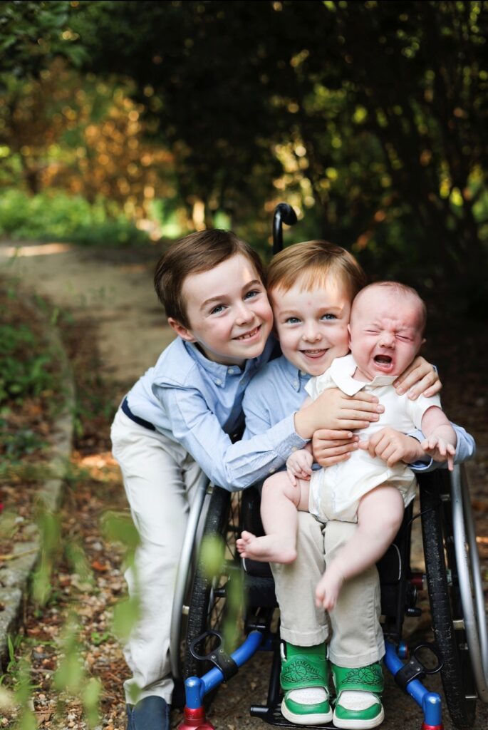 Three young boys outdoors, two smiling and hugging a crying baby. One boy uses a wheelchair, another stands beside him, and the baby sits on their laps. Sunlight filters through the trees as Elliotts Story unfolds in the background.