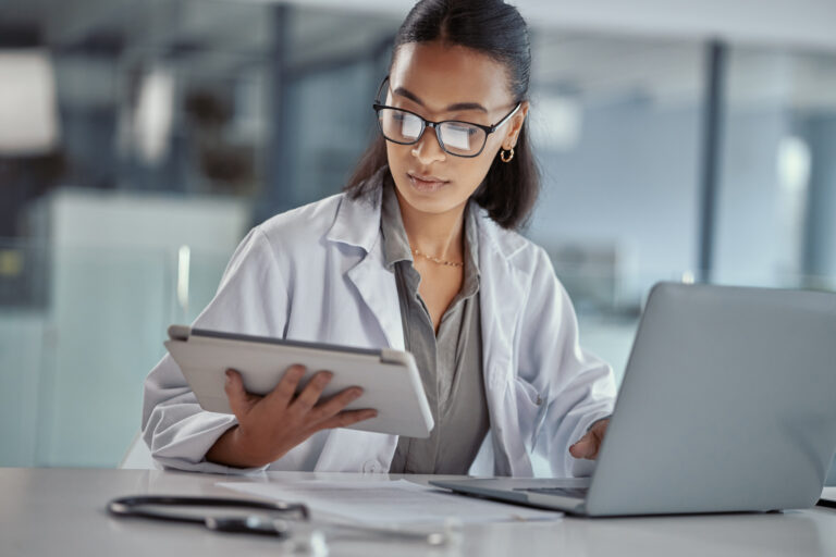 A woman in a lab coat and glasses sits at a desk with a laptop, holding a tablet. Medical tools lie on the desk, and the blurred background suggests a modern Numotion office or lab setting.