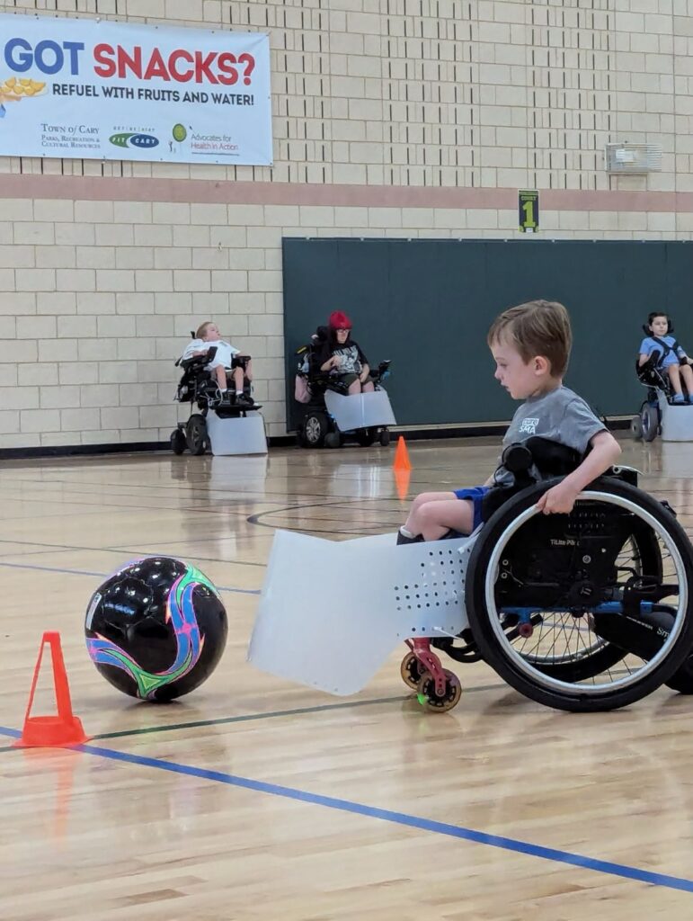A young boy in a wheelchair uses a plastic guard to push a soccer ball in a gym, while other children in wheelchairs participate in the background. Orange cones mark the play area. A banner promotes healthy snacks and highlights Elliotts Story.