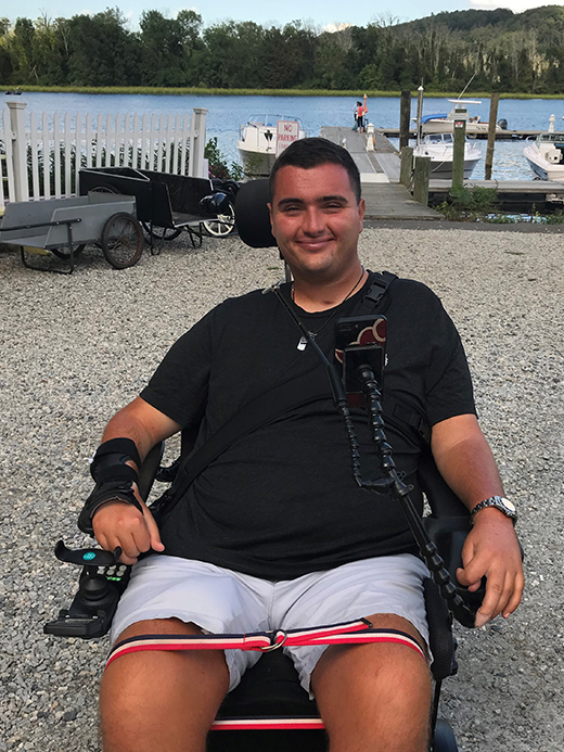 A man sits in a powered wheelchair on a gravel path near a docked marina with boats and water in the background. He is smiling and wearing a black t-shirt and shorts, with trees and hills visible beyond the marina.