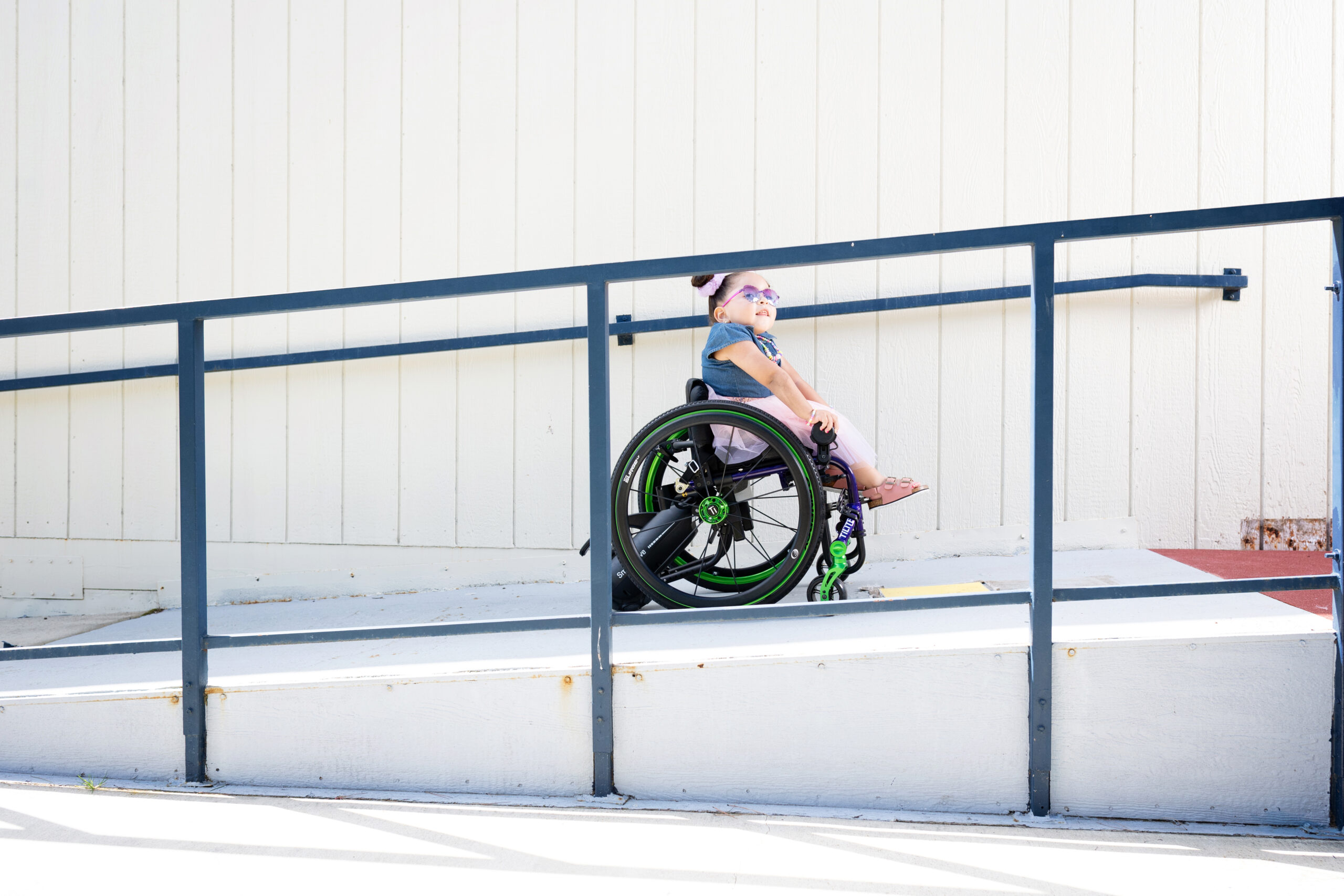 A young pediatric girl uses a wheelchair with a Power Assist add-on as she moves up an outdoor accessibility ramp beside a light-colored building. She wears glasses, a mask, and a blue shirt, looking ahead confidently.