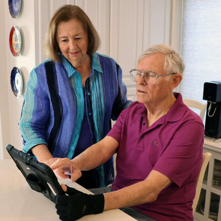 An older man in a purple shirt and black glove uses a tablet with TouchTalk Plus at a table, while an older woman in a blue jacket stands beside him, watching supportively.